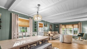 Un espace repas avec une table et des bancs en bois, un lustre et une vue sur l’extérieur grâce à de grandes fenêtres ornées de rideaux.