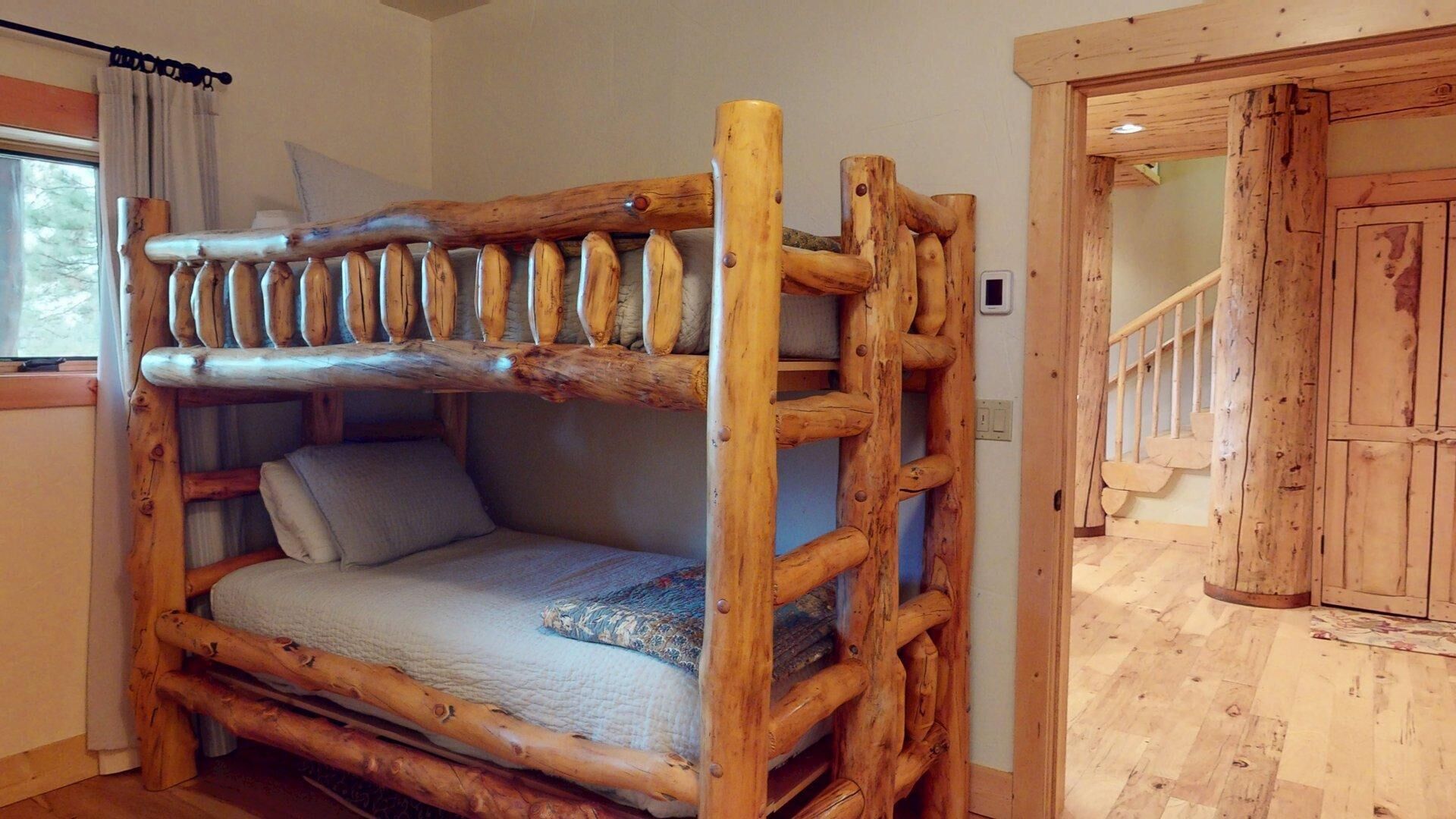 A rustic wooden bunk bed with a simple design, a small window with curtains, and a wooden staircase in the background.