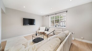 Ein modernes Wohnzimmer mit einer großen Couch, einem Couchtisch und einem an der Wand montierten Fernseher. Ein Fenster bietet einen Ausblick nach draußen.