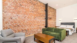 Ein Zimmer mit Backsteinwand, einem grünen Sofa, einem grauen Sessel, einem Holztisch und einem Bett mit weißer Bettwäsche.