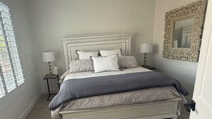Un dormitorio con una cama grande, dos mesitas de noche con lámparas, una ventana con persianas y un espejo con marco decorativo.