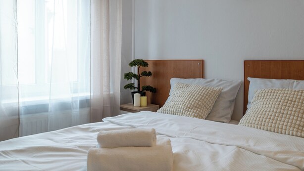 A neatly made bed with white linens and two towels, a bedside table with a small potted plant, and a painting on the wall. - Zabrze City Comfort Room 1 (Zabrze)