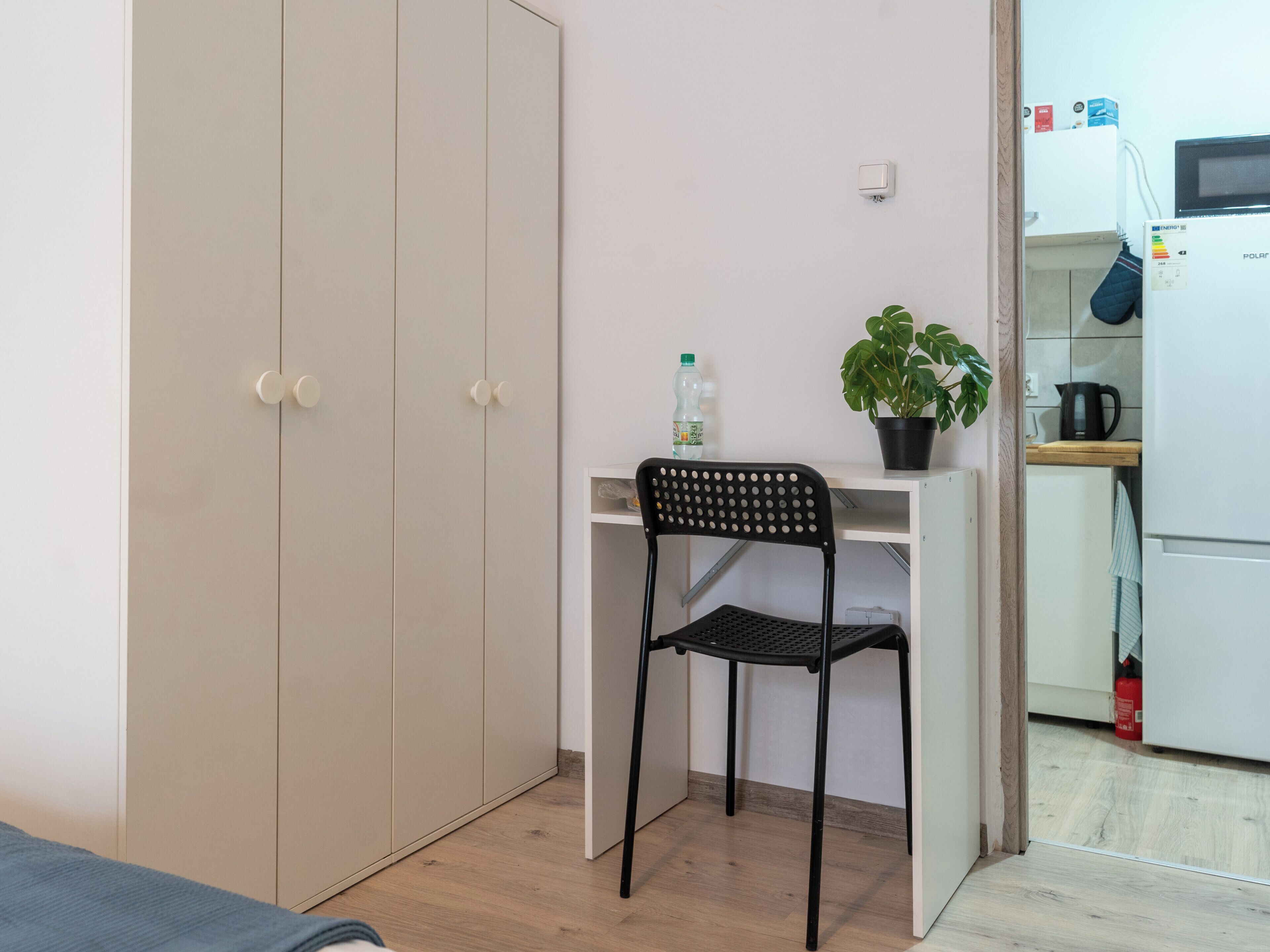 Une petite pièce comprenant une armoire, un bureau avec une chaise, une plante en pot, une bouteille et un four à micro-ondes.