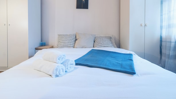 A neatly made bed with white and blue linens, a blue blanket, and rolled towels. There is a checkered pillow and a plain pillow on the bed. A framed picture hangs on the wall above the bed.