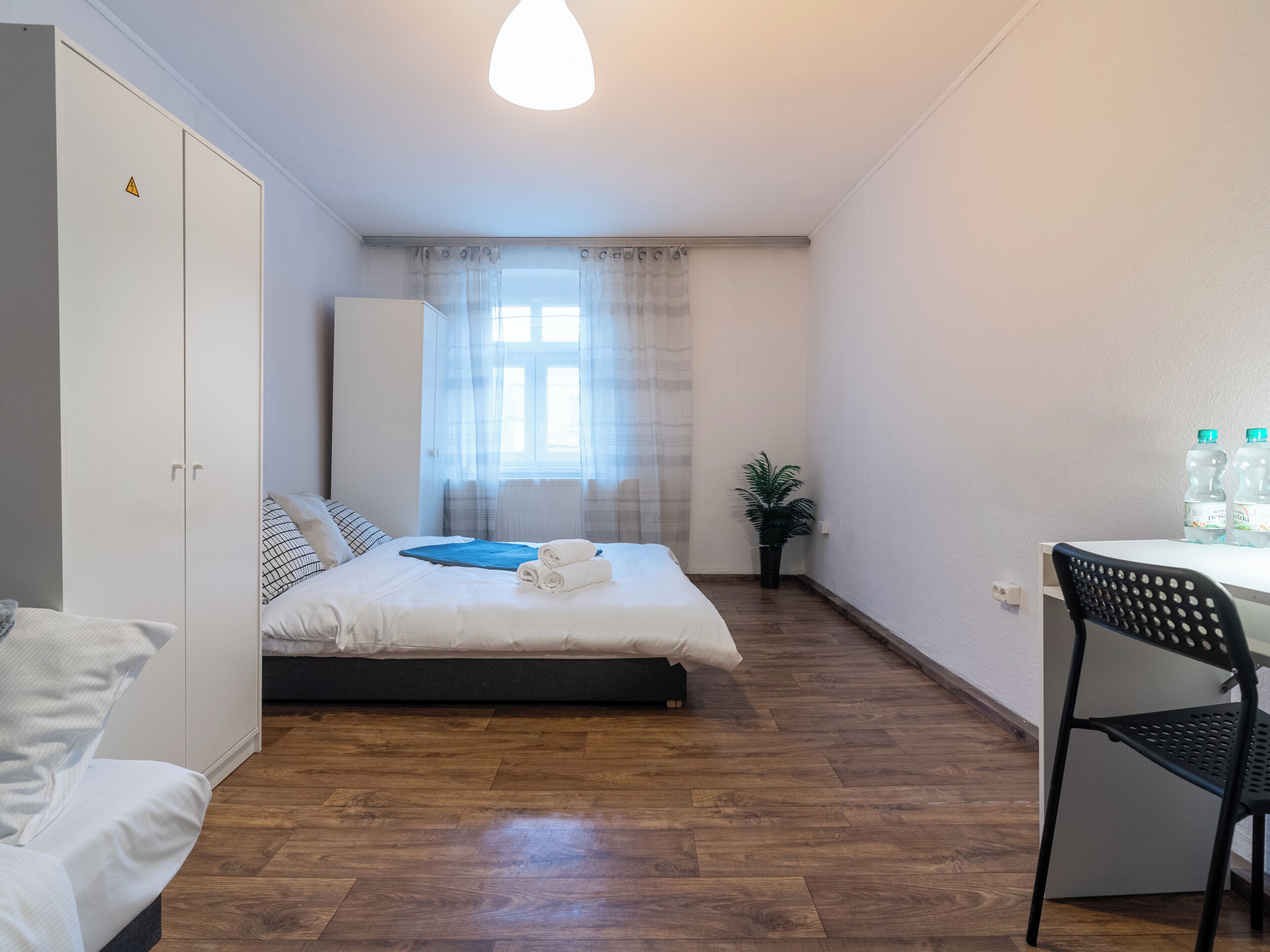 Ein Schlafzimmer mit Bett, Schreibtisch, Stuhl, Kleiderschrank und einem Fenster mit Vorhängen.