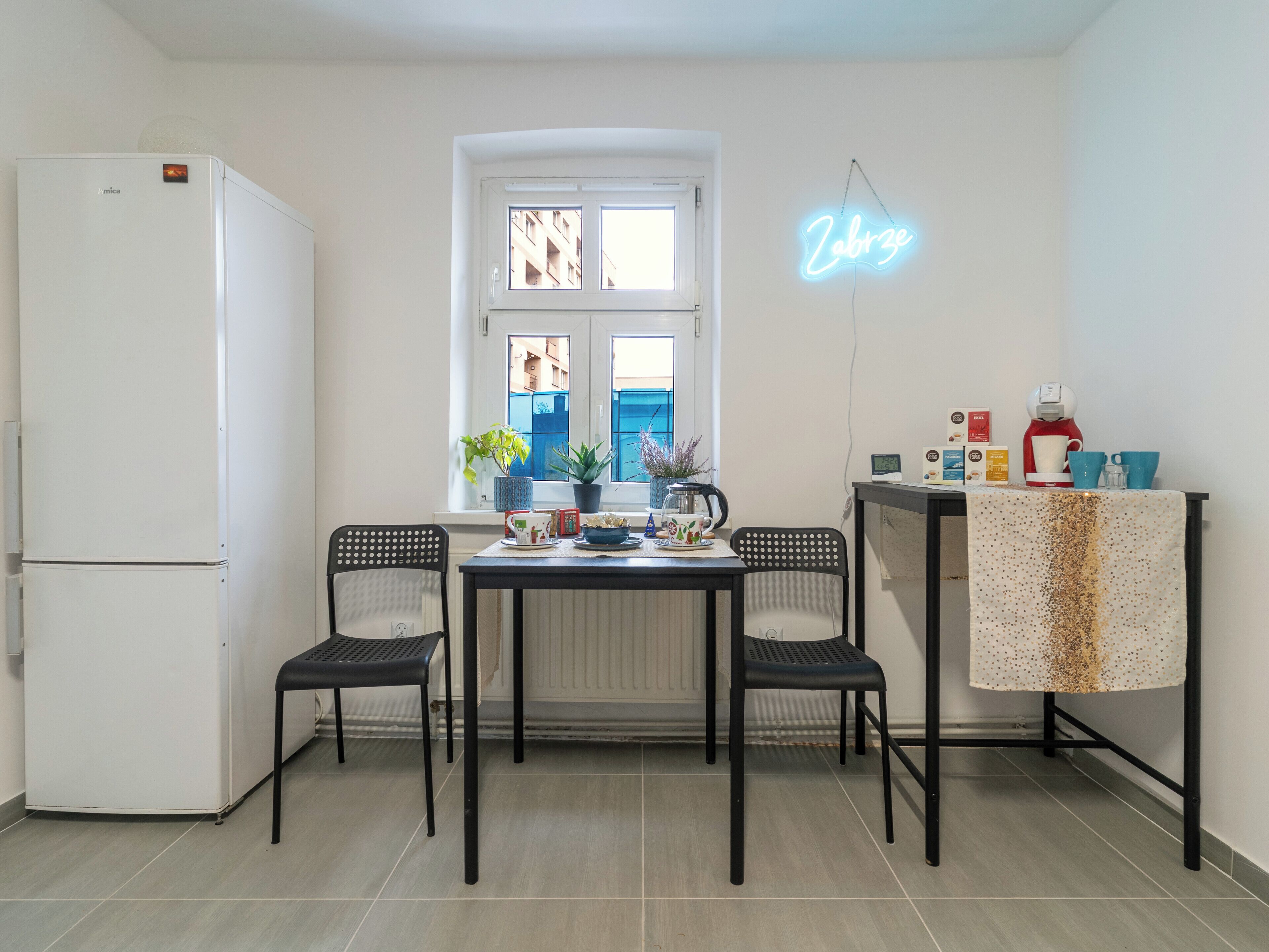 A compact kitchen with a white refrigerator, two black chairs, a dining table, and a neon sign.