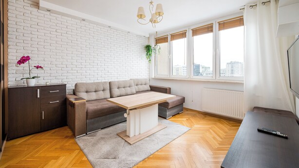 A modern living room with a sectional sofa, a wooden coffee table, a dark cabinet, and a large window with a view of the city. - Solidarności City View Apartment (Warszawa)