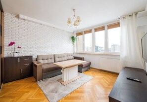 A modern living room with a sectional sofa, a wooden coffee table, a dark cabinet, and a large window with a view of the city. - Solidarności City View Apartment (Warszawa)