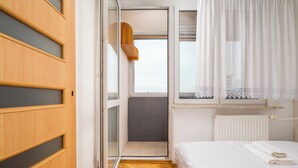 A bedroom with a wooden wardrobe, a bed with white linens, a window with sheer curtains, and a radiator. - Solidarności City View Apartment (Warszawa)