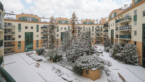 Une cour bordée d’arbres couverts de neige et de bancs, entourée d’immeubles à plusieurs étages dotés de balcons.