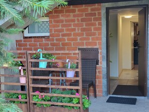 Un mur de briques sur lequel est fixée une étagère en bois, où sont disposées des plantes et des fleurs en pot, une chaise en osier et une porte donnant sur un espace intérieur.