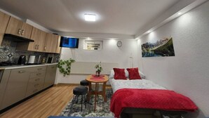 A compact room with a kitchenette, a bed with red bedding, a small table with a plant, and a wall-mounted TV. - Kowary Forest Apartment near Karpacz (Kowary)