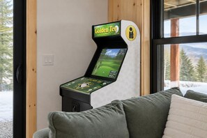 Eine Golf-Videospielkonsole mit dem Markennamen 'Golden Tee' in einem Raum mit Blick auf Bäume und Berge durch ein Fenster.