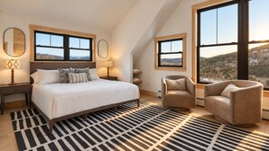 Een slaapkamer met een groot bed, twee nachtkastjes, een stoel en uitzicht op de omgeving door grote ramen.