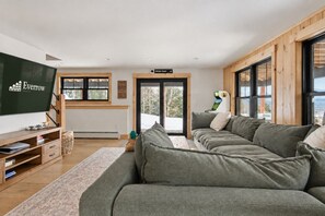 A modern living room with a grey sectional sofa, a flat-screen TV, and a wooden-framed window overlooking a snowy landscape.