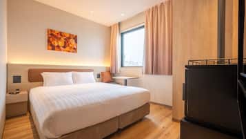 A modern hotel room with a large bed, a small table, a nightstand, a refrigerator, and a window with curtains.
