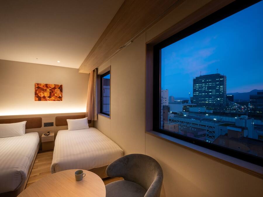 A hotel room with two beds, a small table with a cup, a chair, a painting on the wall, and a large window with a city view.