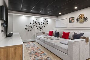 A modern living room with a grey sofa, a coffee table, a TV, and a wall art piece. - Epic House for Groups: 10 Mins to Sunday River (Bethel)