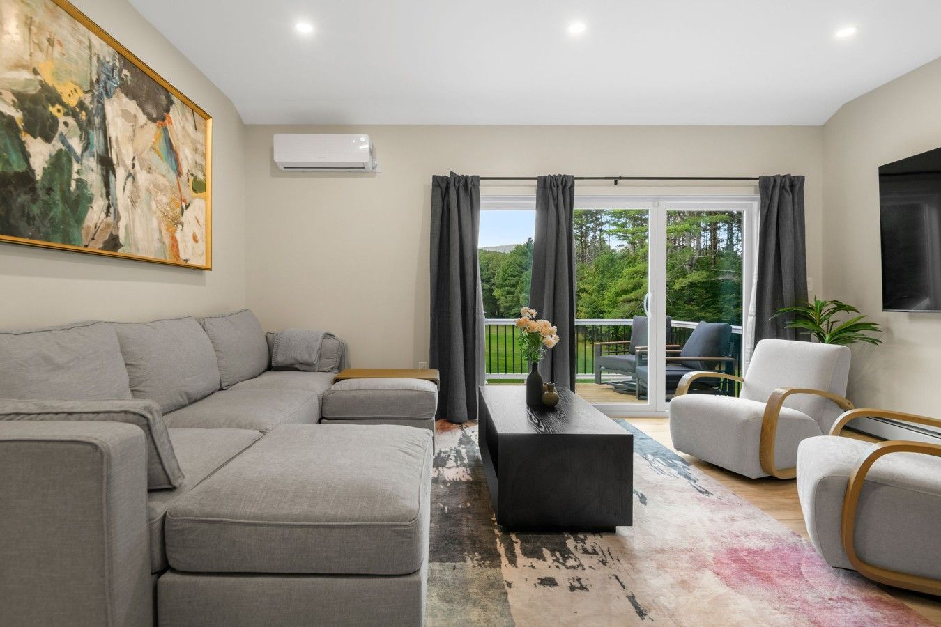 A modern living room with a sectional sofa, a coffee table, and a TV. There is a sliding glass door leading to an outdoor area with a view of trees.