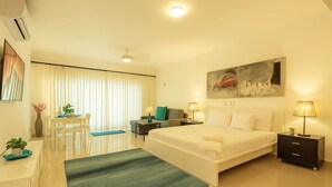A modern hotel room with a large bed, a desk with a chair, a dining table with chairs, and a large window with curtains. - Stunning  Beach Front Condo, King Bed, Stunning Beach and Ocean Views (Cabarete)
