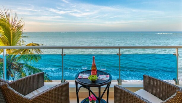 A balcony with wicker furniture, a table with a bottle and glasses, and a view of the ocean. - Stunning  Beach Front Condo, King Bed, Stunning Beach and Ocean Views (Cabarete)