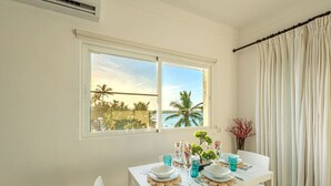 A dining area with a table set for two, a view of palm trees and water through large windows, and a curtain on the right. - Stunning  Beach Front Condo, King Bed, Stunning Beach and Ocean Views (Cabarete)
