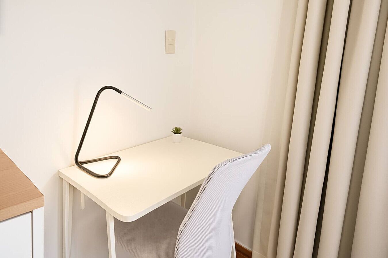 Un petit bureau blanc, agrémenté d’une lampe moderne, d’une petite plante en pot et d’une chaise blanche.