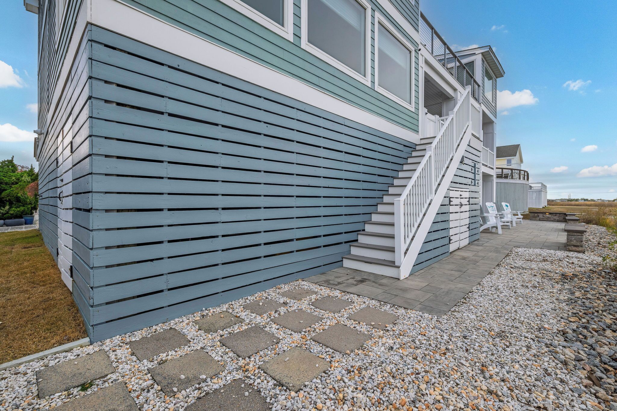 A two-story house with a blue exterior, a stone pathway, and a small patio area with white chairs.