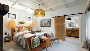Une chambre avec un grand lit, deux chaises en bois, une petite table, un luminaire suspendu, un tableau et une porte coulissante de style grange donnant sur une salle de bain.