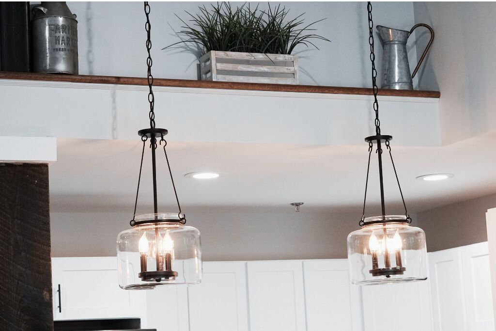 Une cuisine avec des meubles blancs, une étagère en bois ornée d’une plante et deux lampes suspendues.
