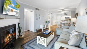 A modern living room with a fireplace, a television, and a dining area with a kitchen in the background. - Ocean's Outside: Relaxing Oceanfront Escape (Kill Devil Hills)