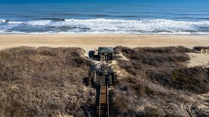 A wooden boardwalk leading to a beach house with a green roof, surrounded by dry vegetation. - Ocean's Outside: Relaxing Oceanfront Escape (Kill Devil Hills)