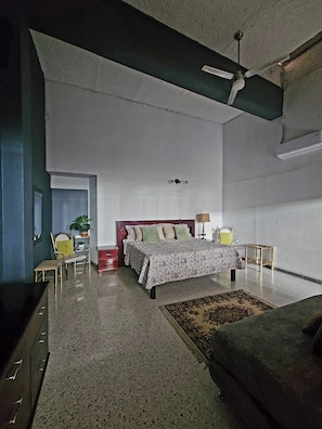 Un dormitorio con cama, mesita de noche, silla, pequeña mesa, sofá, ventilador de techo y aire acondicionado instalado en la pared.