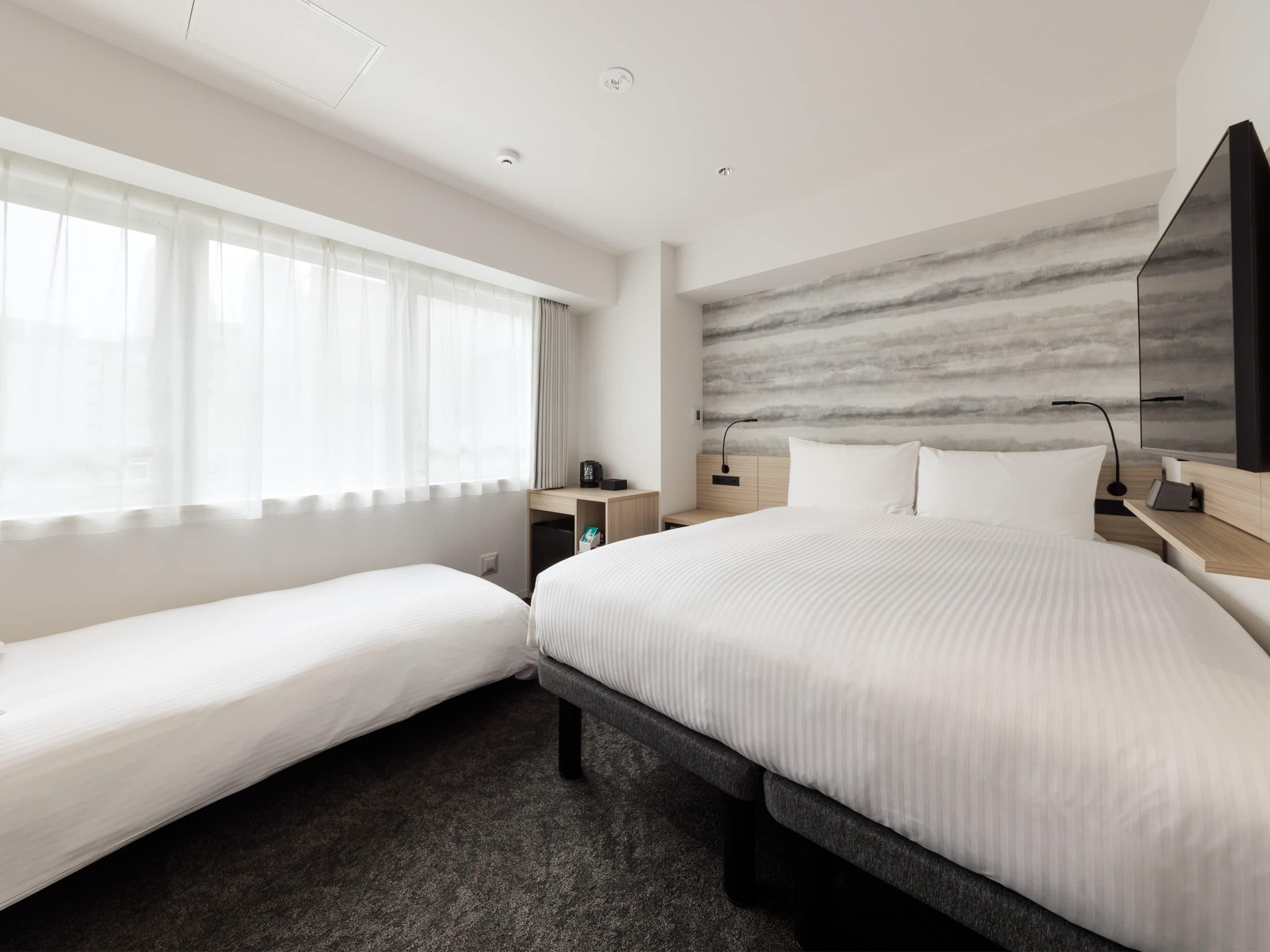 A hotel room with a large bed, a smaller bed, a desk, a chair, a nightstand, a lamp, and a window with sheer curtains.