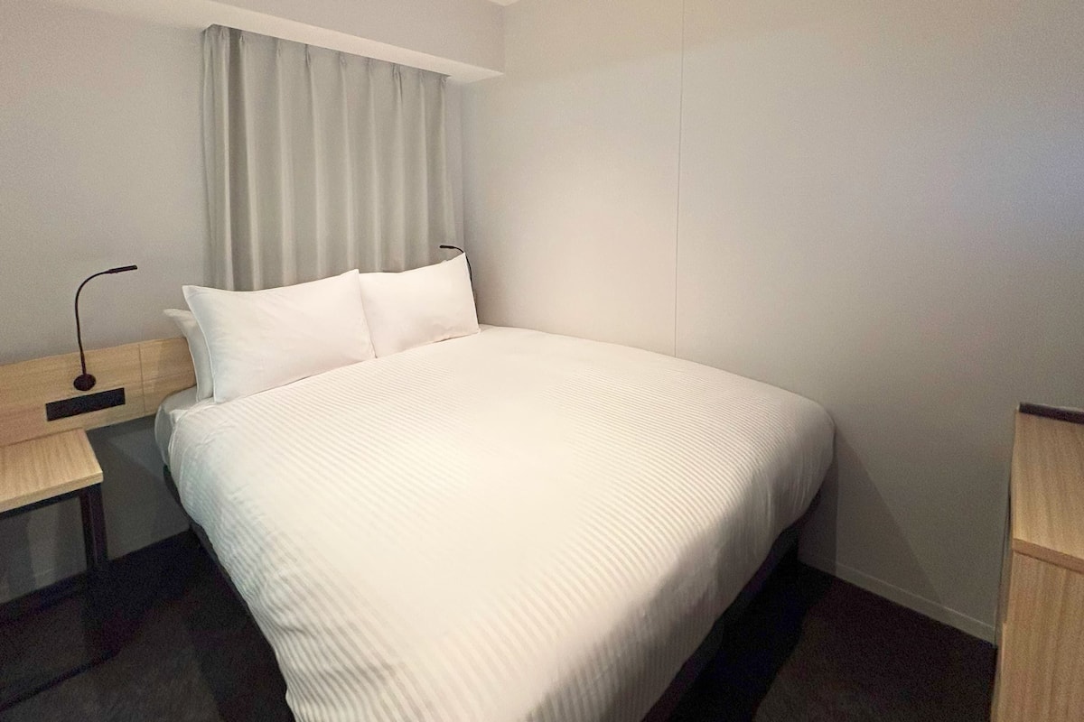 A hotel room with a large bed, a bedside table, a lamp, and a window with curtains.