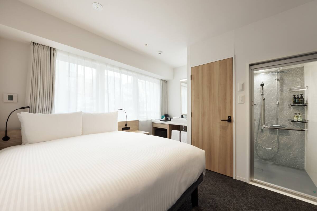 A hotel room with a large bed, a bedside table, a desk, a shower area, and a mirror.