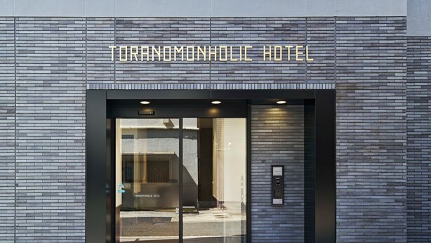 The exterior of Toranomoholic Hotel with a modern brick facade, large glass entrance doors, and a visible elevator. - TORANOMONHOLIC HOTEL (Tokyo)