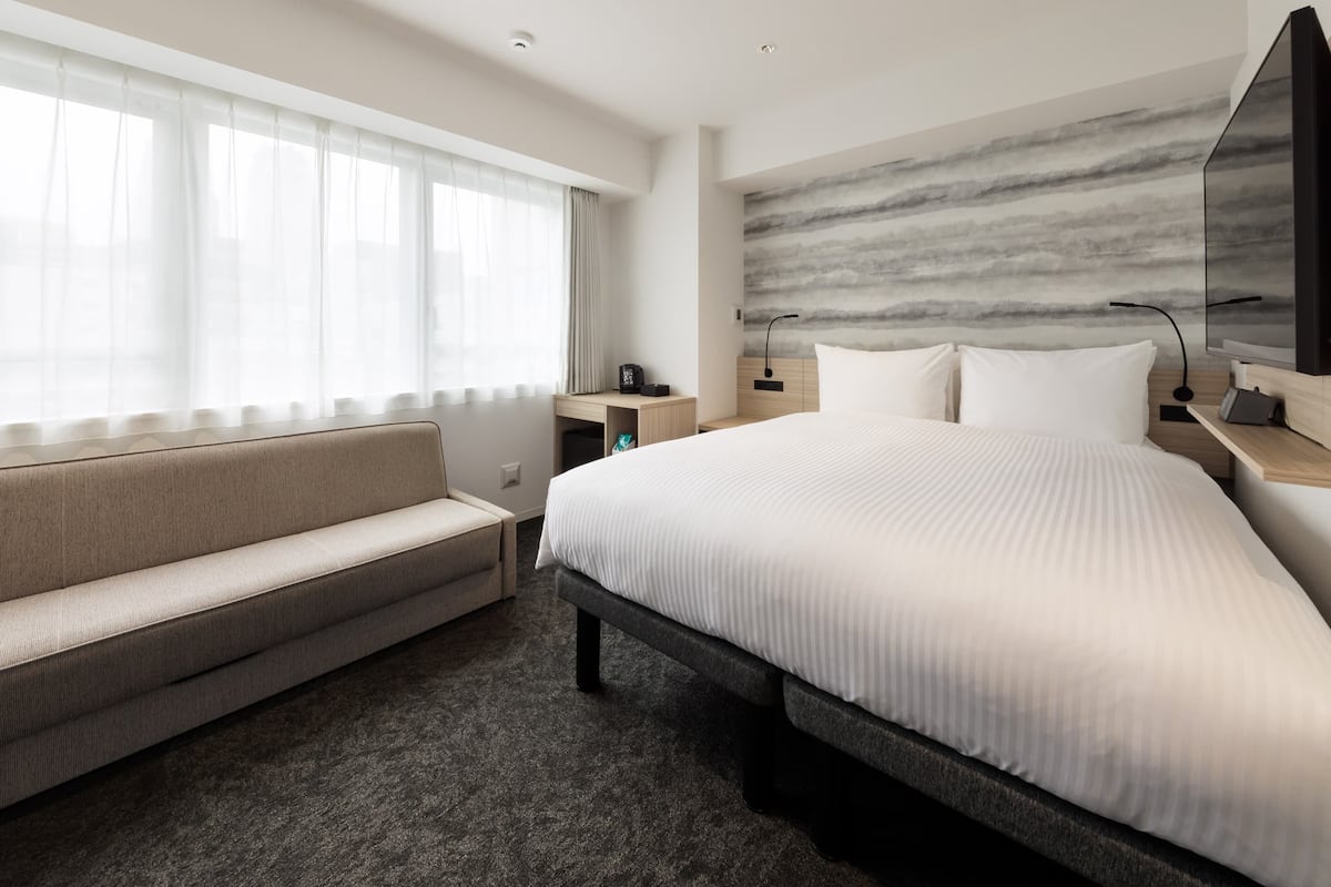 A modern hotel room with a large bed, a sofa, a desk, and a wall-mounted TV.