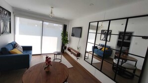 A modern living room with a large sliding glass door, a blue sofa, a wooden coffee table with a vase, and a mirrored wall reflecting the room. - Cozy apartment with ocean view (Lima)