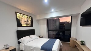 A modern bedroom with a large bed, a bedside table with a lamp, a window with a city view, and a wall-mounted TV. - Cozy apartment with ocean view (Lima)