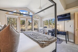 Une chambre avec un grand lit, un bureau, une chaise, une télévision et de grandes fenêtres donnant sur la forêt.