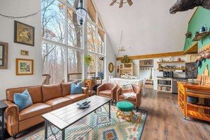 A spacious living room with a large window, a brown leather sofa, a wooden coffee table, and a patterned rug. - Shenandoah Valley Cabin · Hot Tub & Barrel Sauna (Hedgesville)
