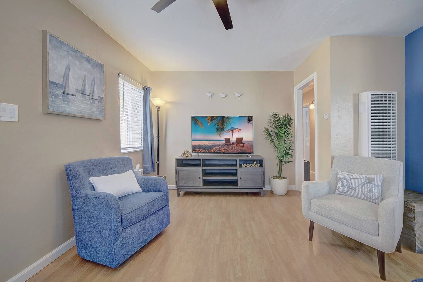 A living room with a blue sofa, a white armchair, a TV on a grey cabinet, a painting, and a potted plant.