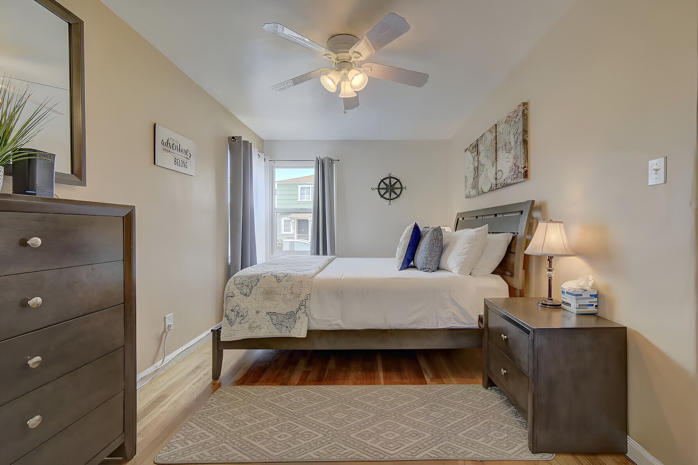 Une chambre à coucher comprenant un lit, une commode, une table de chevet, un ventilateur de plafond et une fenêtre avec des rideaux.