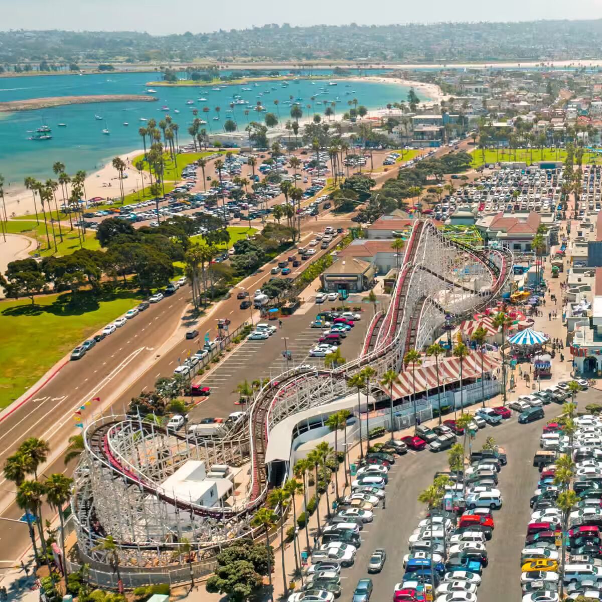 A coastal amusement park with a roller coaster, parking lots, and a beach in the background.