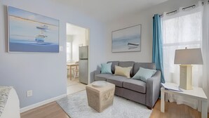 A modern living room with a grey sofa, a small table, and a lamp. There are two paintings on the wall, a window with curtains, and a door leading to another room. - Studio by the Beach • A/C, FREE Bikes & Beach Gear (San Diego)