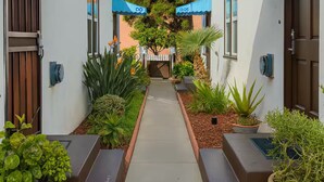 A pathway leading to a row of numbered hotel rooms, each with a door and a small garden area. - Studio by the Beach • A/C, FREE Bikes & Beach Gear (San Diego)