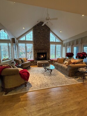 A spacious living room with a stone fireplace, large windows, and a high ceiling fan. - Cozy and elegant lakeside home. (Crossville)
