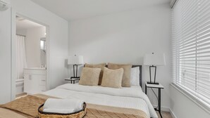 Ein Schlafzimmer mit einem Bett, zwei Nachttischen, einer Lampe auf jedem Tisch, einem Fenster mit Jalousien und einem durch eine offene Tür sichtbaren Badezimmer.