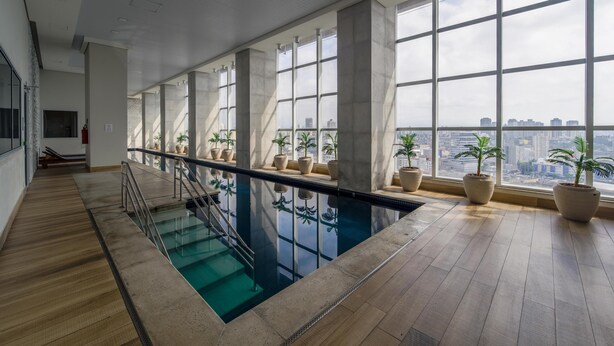 A modern indoor swimming pool with large windows and potted plants. - Modern studio apartment with queen-size bed in Cityplex Osasco. (Osasco)